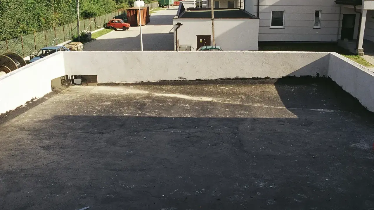 Płaski dach z widocznym odpływem, gotowy na deszcz. W tle budynki i drzewa.
