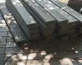 Stos ciemnych desek tarasowych, czyli kantówka kompozytowa, leży na drewnianym podłożu. Cienie liści tworzą na powierzchni wzory.