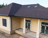 Nowy dom z żółtej cegły, z ciemnobrązowym pokryciem gontem na dachu, ozdobiony białymi kolumnami i oknami dachowymi.