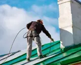 Pracownik w kasku i uprzęży zabezpieczającej maluje zielony stolec dachowy.