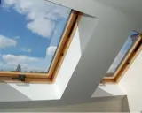 Obróbka blacharska okna dachowego wykonana z drewna, z widokiem na błękitne niebo i chmury.