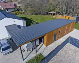 Nowoczesny budynek z drewnianą elewacją i dachem pulpitowym. Obok stoi przykryta plandeką przyczepa i samochód.