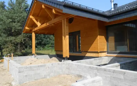 Drewniany dom w budowie, z widocznym fundamentem z bloczków betonowych i dachem o kącie nachylenia 12 stopni.