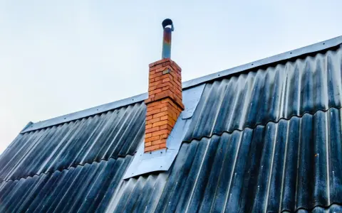 Stary, ceglany komin z metalową rurą wystaje ponad falistą, ciemną blachę dachową. Blacha wokół komina jest przycięta i zabezpieczona.