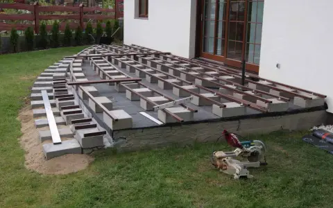 Budowa tarasu: betonowe bloczki i legary tworzą konstrukcję pod deski. Widać piasek, trawę i piłę.