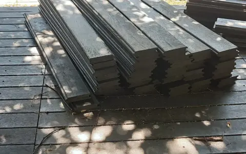 Stos ciemnych desek tarasowych, czyli kantówka kompozytowa, leży na drewnianym podłożu. Cienie liści tworzą na powierzchni wzory.