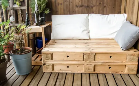 Meble z palet na balkon: wygodna sofa z szufladami, białe i szare poduszki, rośliny w doniczkach, drewniana ściana i podłoga.