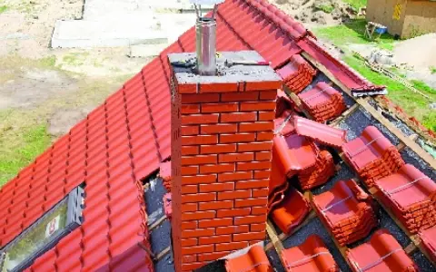 Obróbka komina okrągłego blachą trapezową na czerwonym dachu. Dachówka w paczkach czeka na ułożenie.