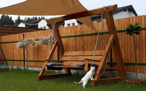 Drewniana huśtawka ogrodowa jak zrobić? Biały kot pilnuje relaksu na łonie natury.
