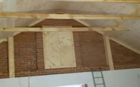 Drewniana konstrukcja dachu nad ceglaną ścianą, z ozdobnym panelem. Stara więźba dachowa czeka na renowację.