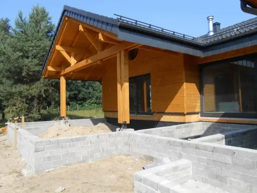 Drewniany dom w budowie, z widocznym fundamentem z bloczków betonowych i dachem o kącie nachylenia 12 stopni.