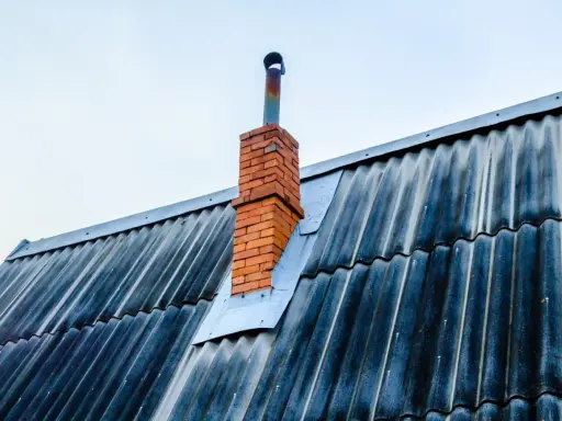 Stary, ceglany komin z metalową rurą wystaje ponad falistą, ciemną blachę dachową. Blacha wokół komina jest przycięta i zabezpieczona.