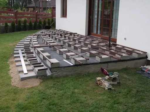 Budowa tarasu: betonowe bloczki i legary tworzą konstrukcję pod deski. Widać piasek, trawę i piłę.