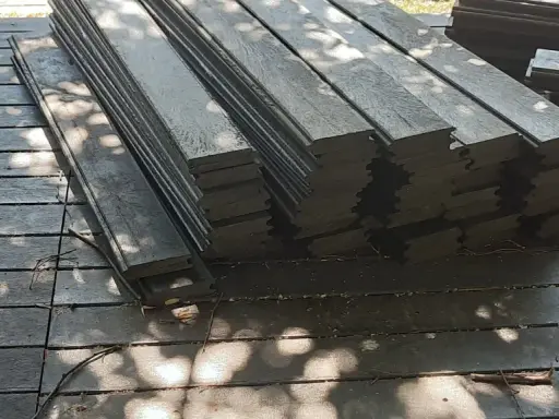 Stos ciemnych desek tarasowych, czyli kantówka kompozytowa, leży na drewnianym podłożu. Cienie liści tworzą na powierzchni wzory.