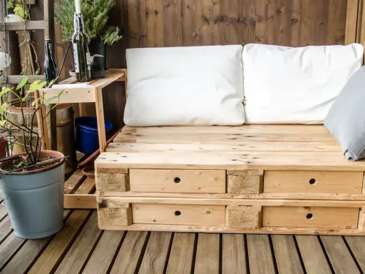 Meble z palet na balkon: wygodna sofa z szufladami, białe i szare poduszki, rośliny w doniczkach, drewniana ściana i podłoga.