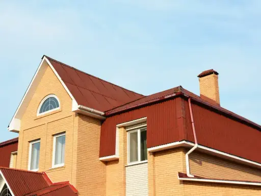 Dach mansardowy konstrukcja z czerwonej blachy falistej, z ceglaną ścianą i kominem.