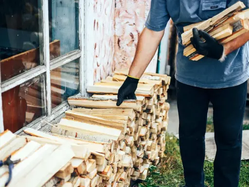 Mężczyzna w rękawiczkach układa drewno przy oknie. Zastanawiasz się, na ile starcza metr drewna na zimę?
