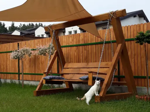 Drewniana huśtawka ogrodowa jak zrobić? Biały kot pilnuje relaksu na łonie natury.