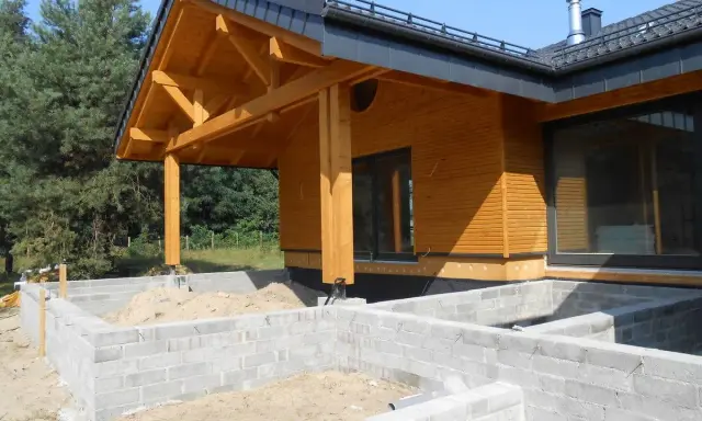 Drewniany dom w budowie, z widocznym fundamentem z bloczków betonowych i dachem o kącie nachylenia 12 stopni.