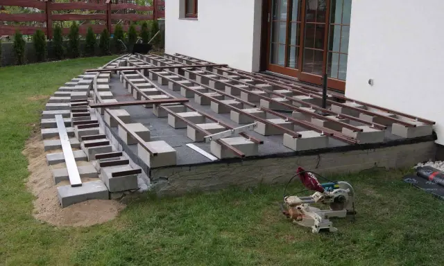Budowa tarasu: betonowe bloczki i legary tworzą konstrukcję pod deski. Widać piasek, trawę i piłę.