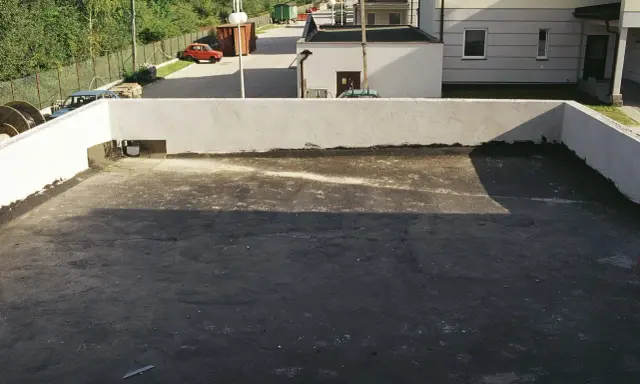Płaski dach z widocznym odpływem, gotowy na deszcz. W tle budynki i drzewa.