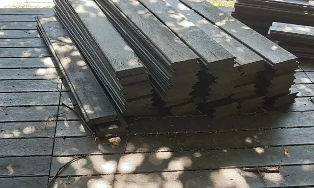Stos ciemnych desek tarasowych, czyli kantówka kompozytowa, leży na drewnianym podłożu. Cienie liści tworzą na powierzchni wzory.