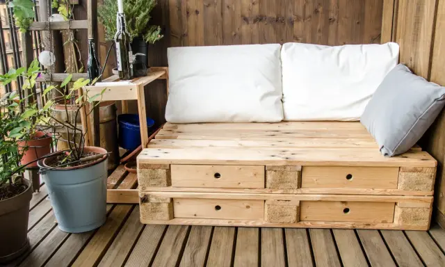 Meble z palet na balkon: wygodna sofa z szufladami, białe i szare poduszki, rośliny w doniczkach, drewniana ściana i podłoga.