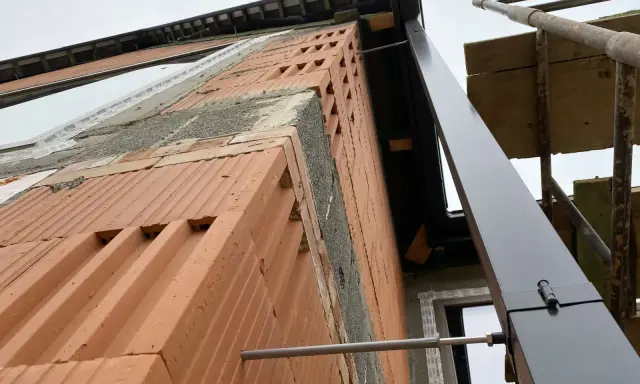 Budowa domu w toku. Widok na ścianę z cegły i rynnę. Trudno ocenić ile rur spustowych na m2 dachu, bo dach jest w budowie.