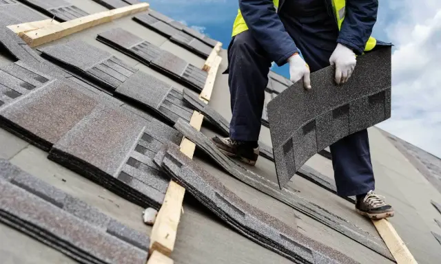 Dachówka bitumiczna układana na dachu. Pracownik w rękawiczkach trzyma kolejny element, przygotowując go do montażu.