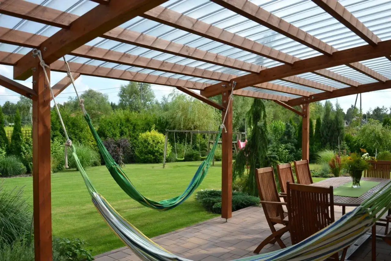 Drewniana pergola z przezroczystym dachem, pod którą wiszą hamaki. Obok stołu z krzesłami, idealne miejsce na relaks. Kantówka kompozytowa zapewnia trwałość konstrukcji.