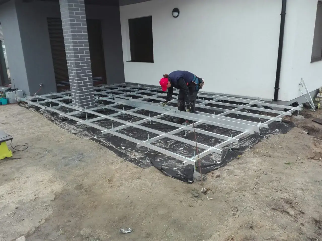 Pracownik buduje taras z deski kompozytowej, montaż bez legarów. Konstrukcja aluminiowa na podłożu.